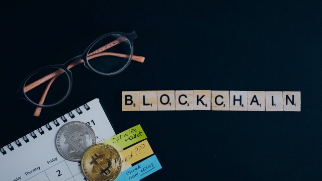 Uma mesa preta com um óculos de armação castanha, um calendário branco com algumas anotações coloridas, duas moedas Bitcoin e alguns blocos de madeira com letras, formando a palavra “blockchain”.