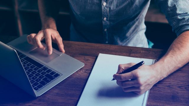 A foto mostra um homem mexendo em um notebook enquanto faz anotações em um bloco de papel