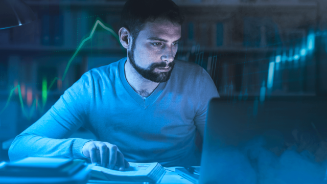 A foto mostra um homem em frente a um notebook com a mão sobre um livro