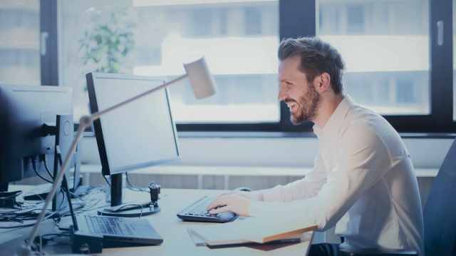 A foto mostra homem sorrindo sentado em uma cadeira utilizando uma computador, que está em uma mesa com um notebook, cadernos e uma luminária, em alusão ao dia a dia de viver de dividendos.