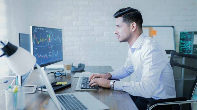 A imagem mostra um homem sentado em uma cadeira utilizando um notebook, como um trader profissional. Ele está vestindo uma camisa social branca e um relógio prateado. Além disso, há vários objetos em sua mesa, como caderno, caneta, óculos, fone de ouvido, café e outros.