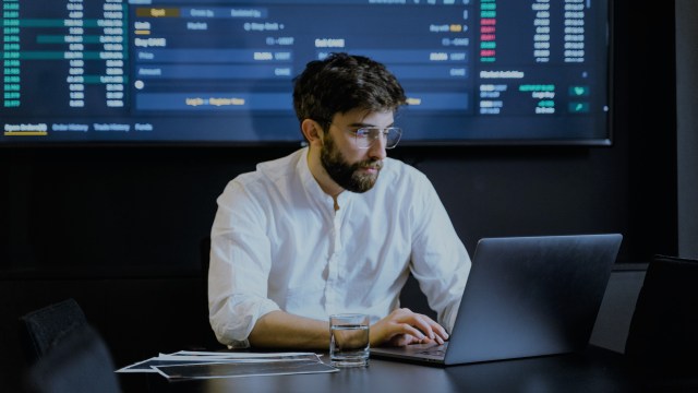 A imagem mostra um homem de óculos e camisa branca mexendo em um computador. Atrás vemos uma tela grande com índices e cotações de ações.