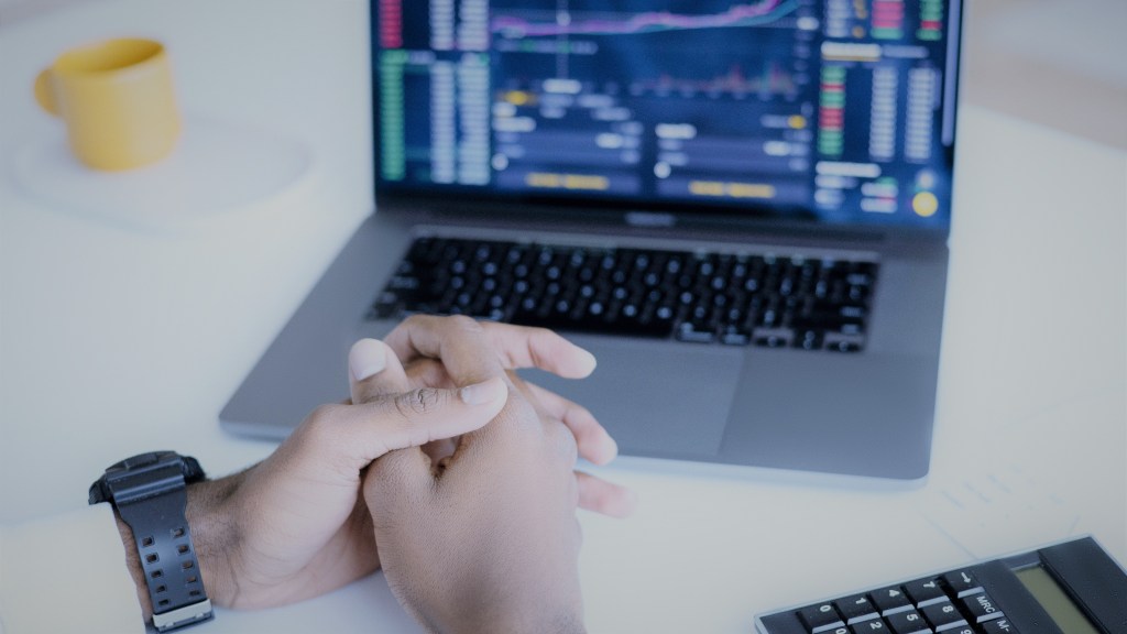 A foto mostra as mãos de um homem cruzadas sobre uma mesa, e à sua frente tem um notebook ligado em uma plataforma para negociar small caps.