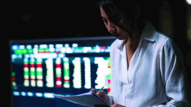Profissional analisa documentos na frente de telas com gráficos de volatilidade da bolsa de valores.