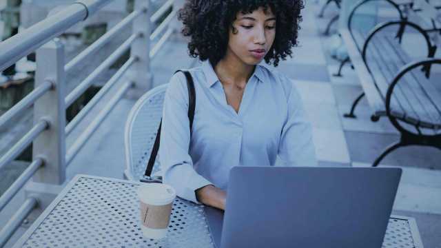 A foto mostra uma mulher, sentada ao ar livre, em uma mesa. Há um copo de café e notebook à sua frente, no qual ela pesquisa sobre como investir em renda variável.