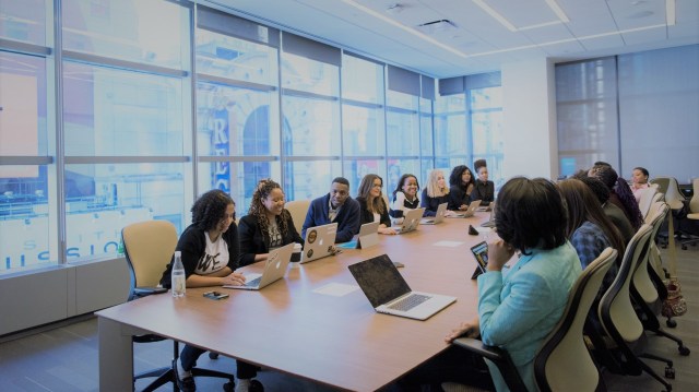 Na imagem existe um grupo de pessoas, majoritariamente negras e pretas, sentadas ao redor de uma mesa retangular participando de uma reunião de empresa. Em cima da mesa temos computadores desktops e garrafas d'água. Ao fundo temos parede com vidros trazendo claridade para a sala.