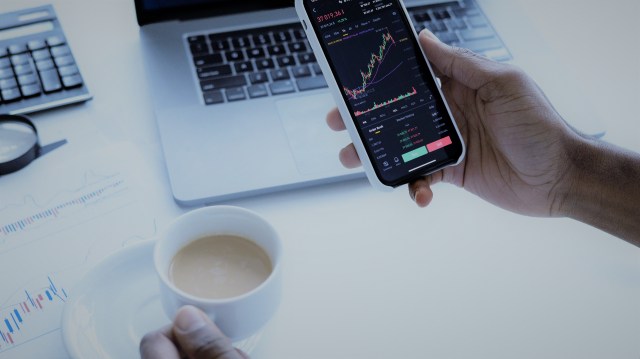 A foto mostra as mãos de uma pessoa. Em uma delas, há uma xícara com café e, na outra, um celular ligado em uma plataforma de investimentos. Ao fundo, há um notebook sobre uma mesa.