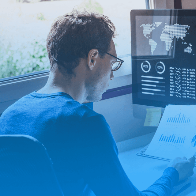 Foto de um homem sentado em frente a um monitor analisando um papel que está em sua mão