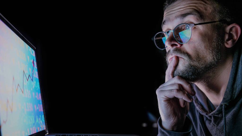 A imagem mostra uma homem branco de óculos e barba com a mão no queixo olhando pensativo para a tela de um notebook que mostra um gráfico de ações em referência a escolha das melhores ações para day trade.