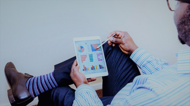 A foto mostra um homem com uma das pernas cruzada, segurando um tablet, no qual há vários gráficos, em referência aos índices das bolsas mundiais.