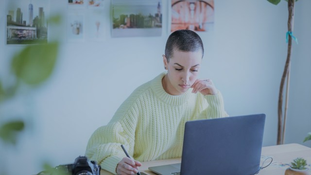 A foto mostra uma mulher sentada à mesa em frente a um notebook, anotando algo em um bloco de anotações ao seu lado.