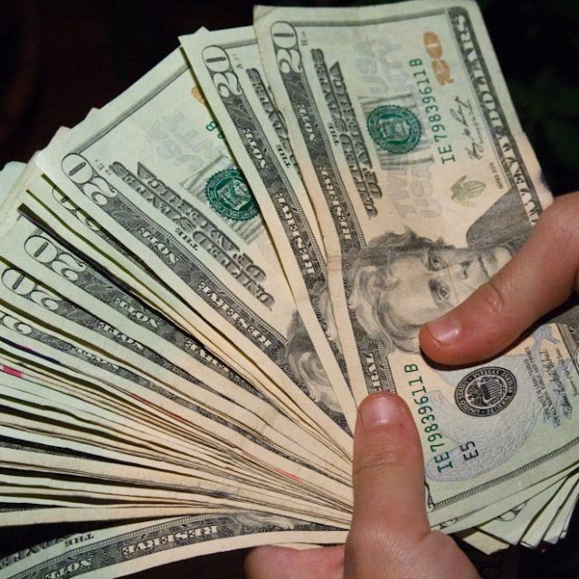 A foto mostra duas mãos segurando notas de dólares em leque