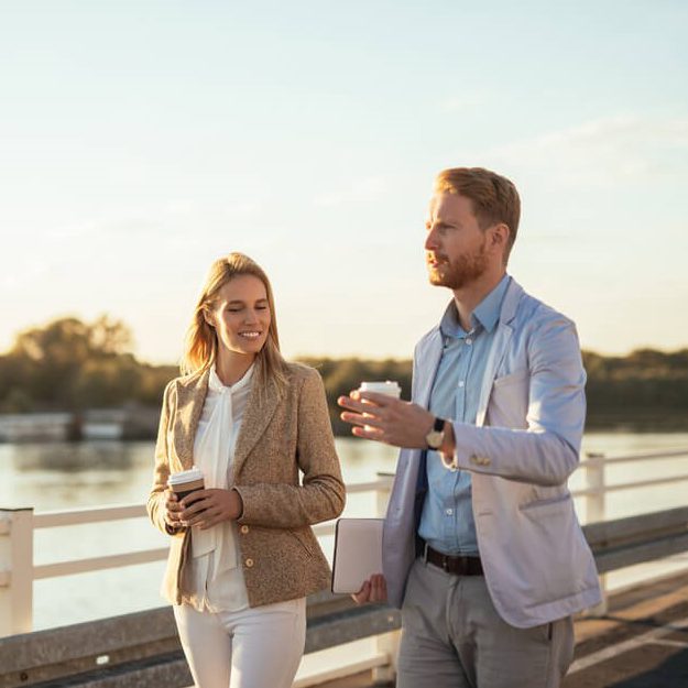 A foto mostra um homem e uma mulher caminhando lado a lado na beira de um lago. Ambos vestem roupas mais formais.