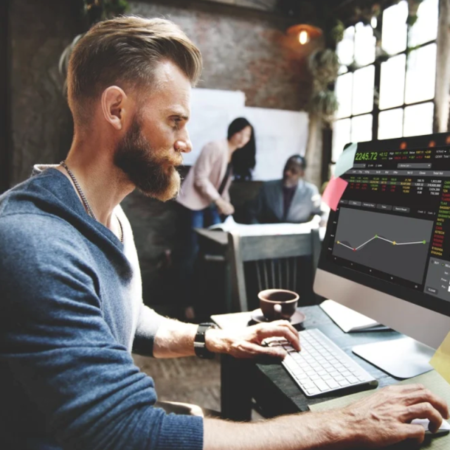 A foto mostra uma homem em um escritório mexendo em seu notebook, que está ligado em uma plataforma de investimentos