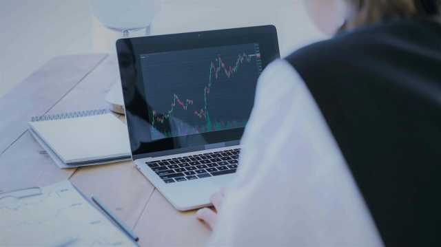 A foto mostra zoom em uma tela de notebook e uma mulher de costas. Na tela do notebook, vemos gráficos em uma plataforma de investimentos, em referência às negociações no mercado de capitais.