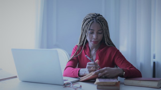 A foto mostra uma mulher em uma sala ou escritório, escrevendo sobre taxa de corretagem em um pequeno cardeno. Na mesa à sua frente, existe um notebook.