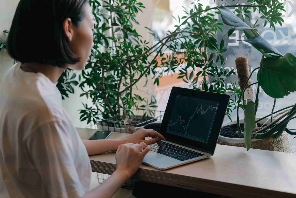 A foto mostra uma mulher de lado para a câmera, à frente de uma mesa de um estabelecimento, com o notebook aberto em uma página de plataforma de investimentos, em referência ao contrato a termo.