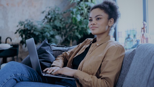 A foto mostra uma mulher de cabelos crespos sentada em um sofá, com um notebook no colo.