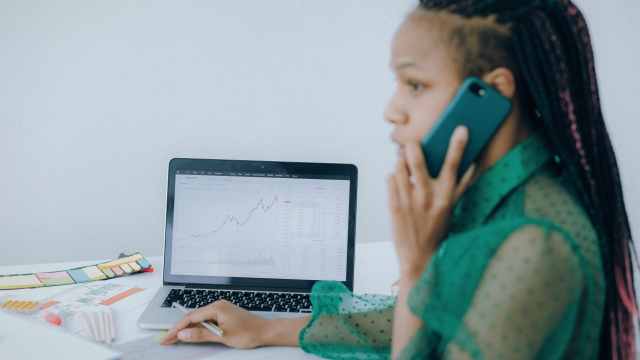 A imagem mostra uma mulher negra de camisa verde escuro e cabelo com dreads atendendo o celular. Ela está com uma caneta na mão e de frente para uma mesa com um notebook que mostra um gráfico dos padrões de candle, em referência a análise do engolfo de baixa.