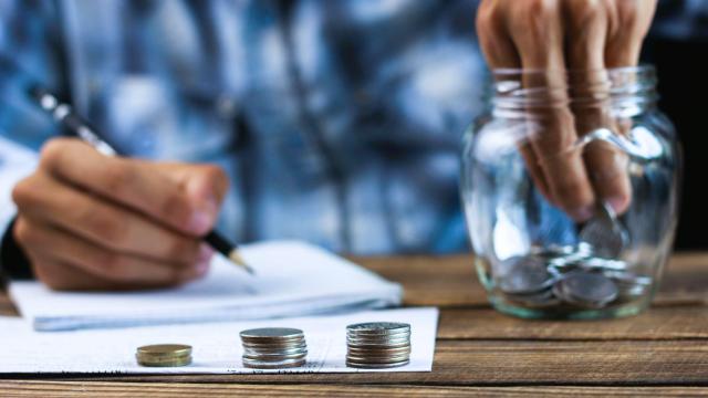 Pessoa fazendo anotações em um caderno enquanto coloca moedas em um pote de vidro, ao lado de pilhas de moedas organizadas sobre a mesa de trabalho, representando o investimento em Tesouro Direto.