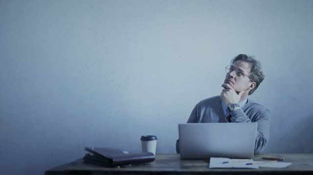 A foto mostra um homem sentado, utilizando um computador em uma mesa, com uma mão no queixo olhando para cima com olhar "pensativo" em referência a questão "como começar a investir na bolsa de valores?".