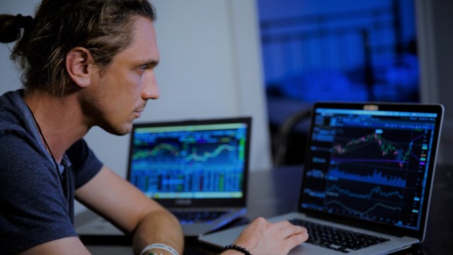 Na imagem vemos um homem branco concentrado mexendo em um computador notebook e também há outro notebook do lado. Nas telas dos dispositivos é possível ver gráficos com linhas coloridas em crescimento e declínio em referência ao processo de comprar ações.