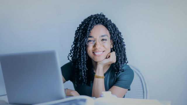 Mulher sentada em uma cadeira com uma mesa à sua frente sobre a qual há um notebook. Ela está sorridente e com a cabeça apoiada em sua mão, olhando diretamente para quem observa a foto.