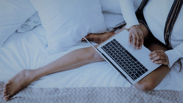 Uma mulher sentada à cama com uma das pernas esticadas mexe no notebook que está em seu colo.