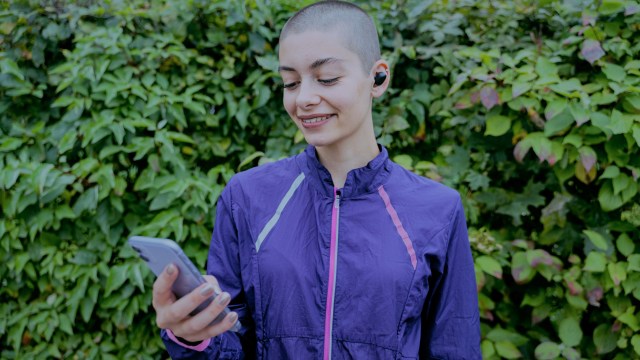 A foto mostra uma mulher em frente a uma parede de folhas segurando um celular com expressão feliz.