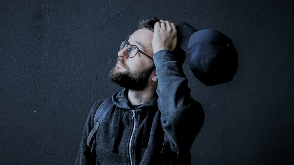 A foto mostra um homem em frente a um fundo preto olhando para cima, segurando um boné com a mão que leva à cabeça, em expressão pensativa.