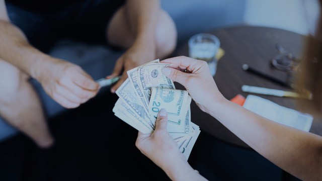A foto mostra as mãos de uma mulher segurando várias notas de dólares. Ao fundo, um homem no sofá e uma mesinha com objetos em cima em referência ao o que é liquidez.