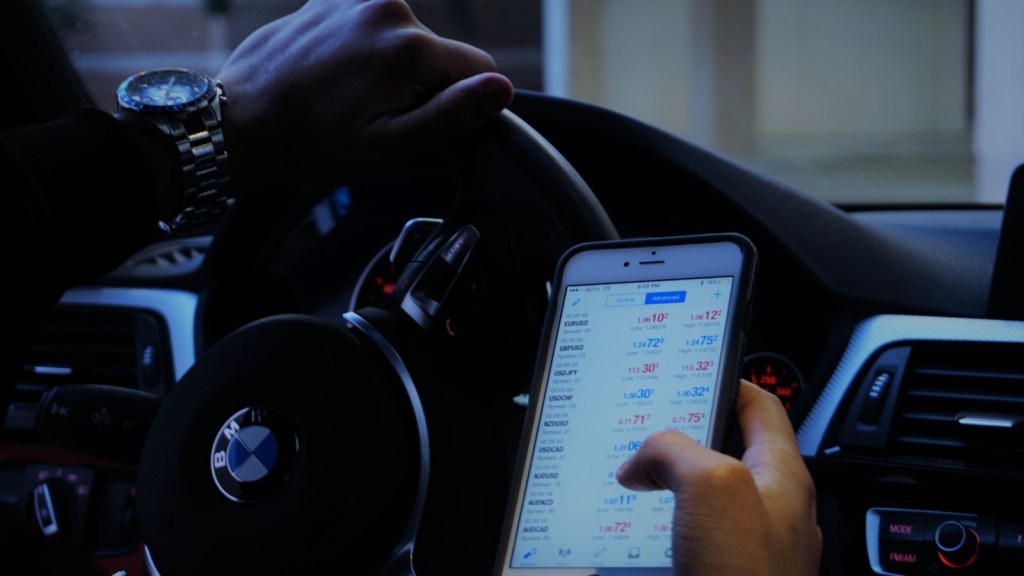 A foto mostra uma pessoa dentro de um carro, com uma das mãos no volante, mexendo no celular, que está ligado na cotação de ações, para acompanhar a volatilidade.