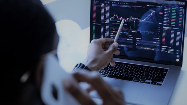 A foto mostra um notebook ligado em uma plataforma de negociações. Em desfoque, de costas para a câmera, um homem aponta para a volatilidade das ações enquanto fala no celular.