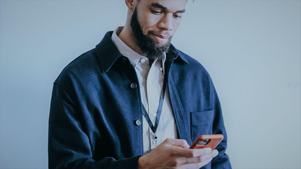 A foto mostra um homem em frente a um fundo branco, olhando para um celular que está em sua mão.