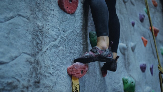 A foto mostra zoom nos pés de uma mulher subindo em uma parede de escalada.