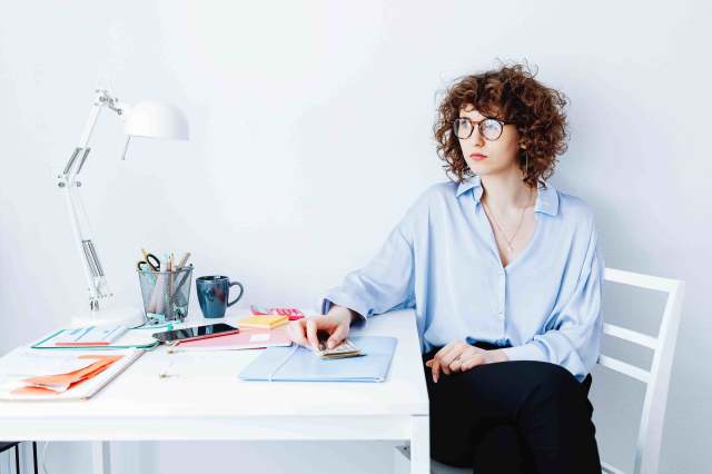 Mulher de cabelo curto cacheado e [oculos, vestindo uma camisa azul clara e calça preta. Ela está sentada em uma cadeira em sua escrivaninha olhando para o lado pensativa em relação aos vieses cognitivos do trader. Na mesa vemos uma luminária, folhas, pasta, porta lápis, dinheiro, calculadora e um celular.