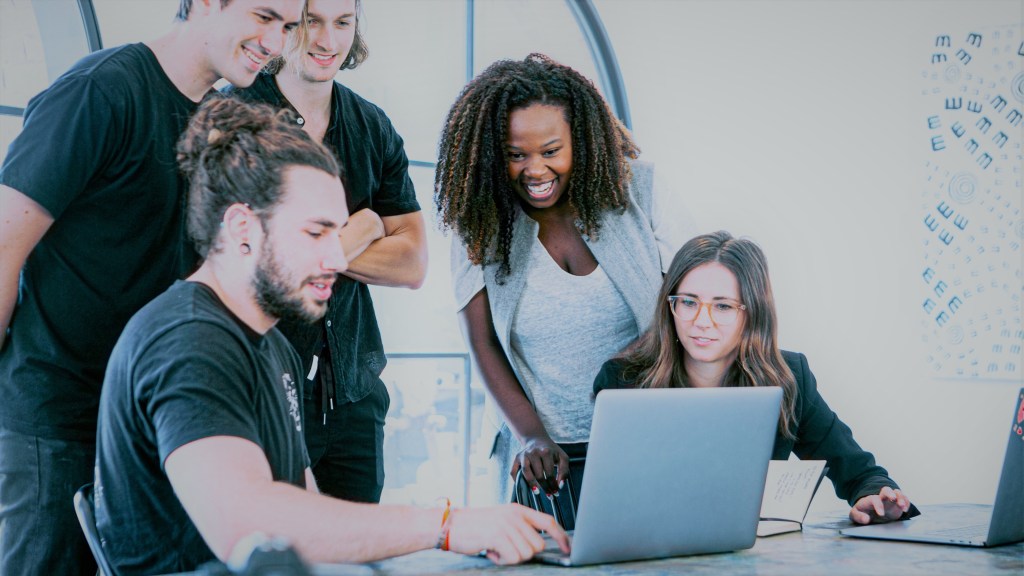 A foto mostra um grupo de cinco pessoas, duas mulheres e três homens, ao redor de uma mesa, em um ambiente de trabalho, olhando para um laptop sobre a mesa.