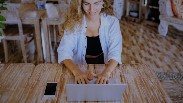 Mulher branca sorrindo e usando laptop em mesa de madeira com celular ao lado, aprendendo como investir em opções de ações, reforçando a importância da pesquisa e análise aprofundada antes de tomar decisões.