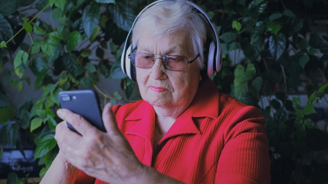 Mulher idosa, com uma camisa vermelha, usando smartphone com fones de ouvido, representando educação financeira acessível, mas também os riscos de investir em opções e a importância de estudar antes de começar a investir.
