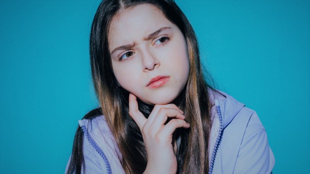 A foto mostra uma menina, em frente a um fundo azul, com uma mão no queixo olhando para o lado, com uma expressão de pensamento.