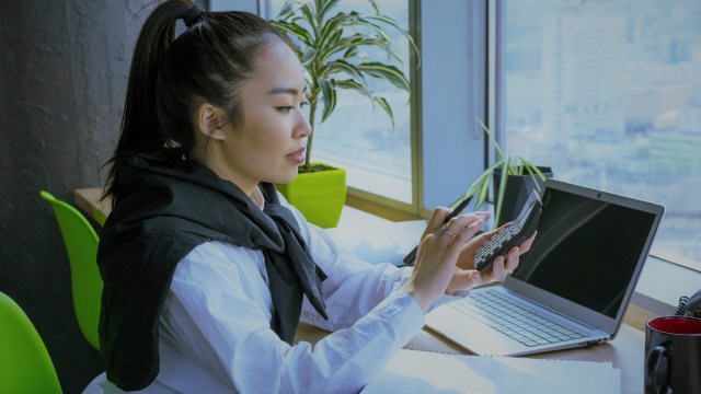 A foto mostra uma mulher sentada em uma cadeira com os braços apoiados em uma mesa, com uma calculadora nas mãos fazendo contas. A sua frente, há um notebook, papéis e uma xícara.