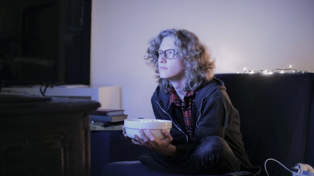 Uma foto de um homem sentado no sofá segurando uma tigela, inclinado para a frente olhando a TV, em referência aos filmes sobre mercado financeiro.