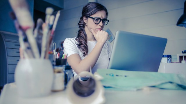 A imagem mostra uma mulher olhando com uma cara de dúvida para o seu notebook. Ela está sentada em uma mesa repleta de materiais de escritório, como canetas e papéis.