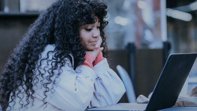 A foto mostra uma mulher de cabelos longos e enrolados sentada à frente de uma mesa, na qual há um notebook aberto. A mulher apoia o rosto sobre ambas as mãos e olha para o notebook.