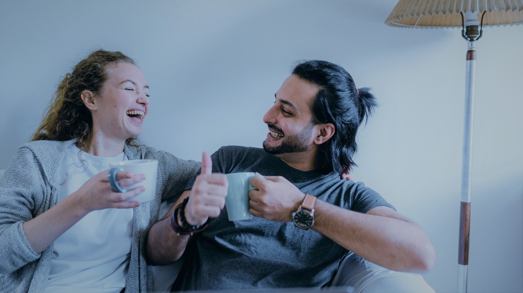 A foto mostra uma mulher e um homem sentados em um sofá. Ambos seguram uma xícara e sorriem um para o outro. O homem faz um sinal de joia, com um dos dedos levantados para a mulher.