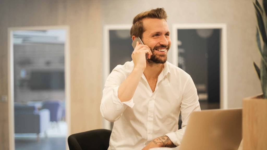 A imagem mostra um homem de barba com uma camisa social branca atendendo o telefone. Em sua frente tem um notebook para fazer sua declaração do imposto de renda. 