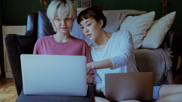 A foto mostra duas mulheres sentadas no chão de uma sala. Ambas têm um notebook no colo, e a mulher que está à direita aponta algo no notebook da outra, em referência às pesquisas sobre o que é inflação.