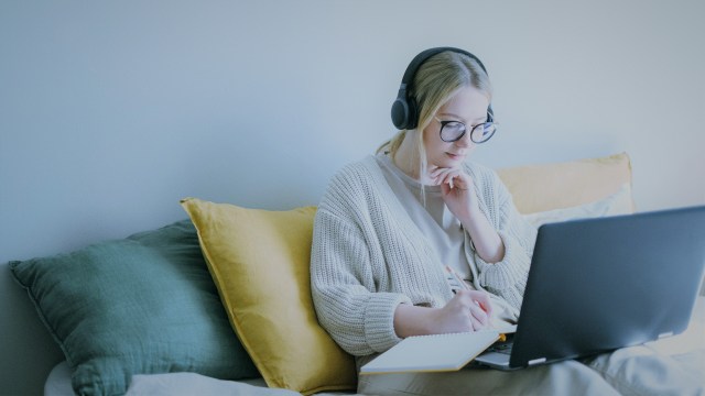 A foto mostra uma mulher loira, sentada em um sofá com várias almofadas. Ela veste um fone e tem um notebook e um caderno no colo, e está escrevendo algo no caderno, em referência ao conhecimento sobre yoy.