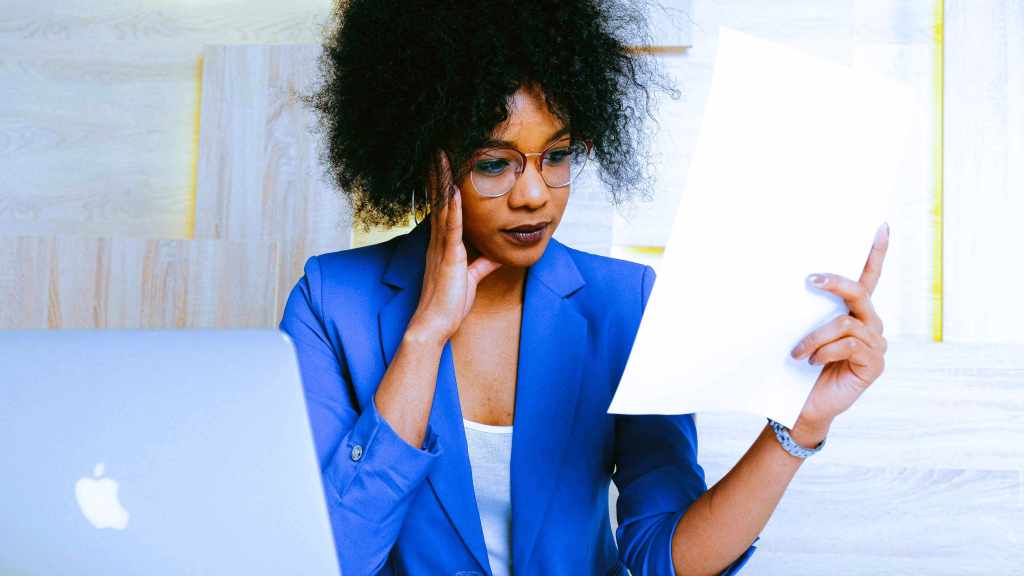 A imagem mostra um mulher negra segurando uma folha de papel com olhar de atenção, em referência ao processo de como declarar ações no Imposto de Renda. Ela também está de frente para um notebook, onde realizará o processo da declaração.