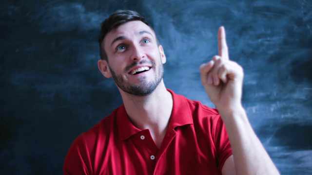 A imagem mostra um homem de cabelo e barba curta, vestindo uma camiseta polo vermelha. Ele está sorrindo e apontando com o dedo indicador para cima, em referência a vida de um trader de sucesso, demonstrando uma expressão de bem-sucedido.